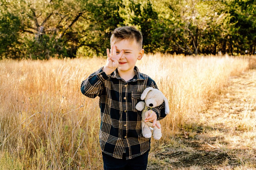 A person in a plaid shirt stands in a golden field holding a stuffed animal toy at sunset.