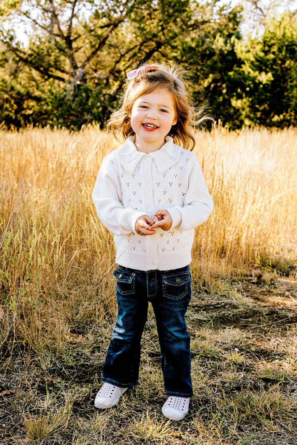 Small child wearing white sweater and jeans stands joyfully in golden autumn field during sunset.