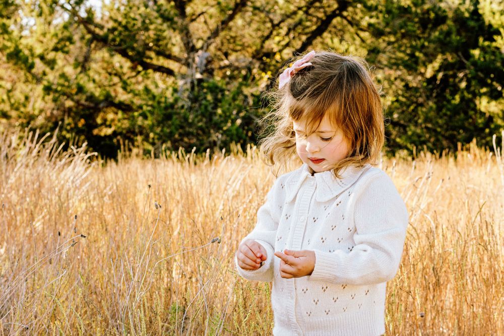 A small child in a white outfit explores a sunlit meadow of golden grasses on an autumn day.