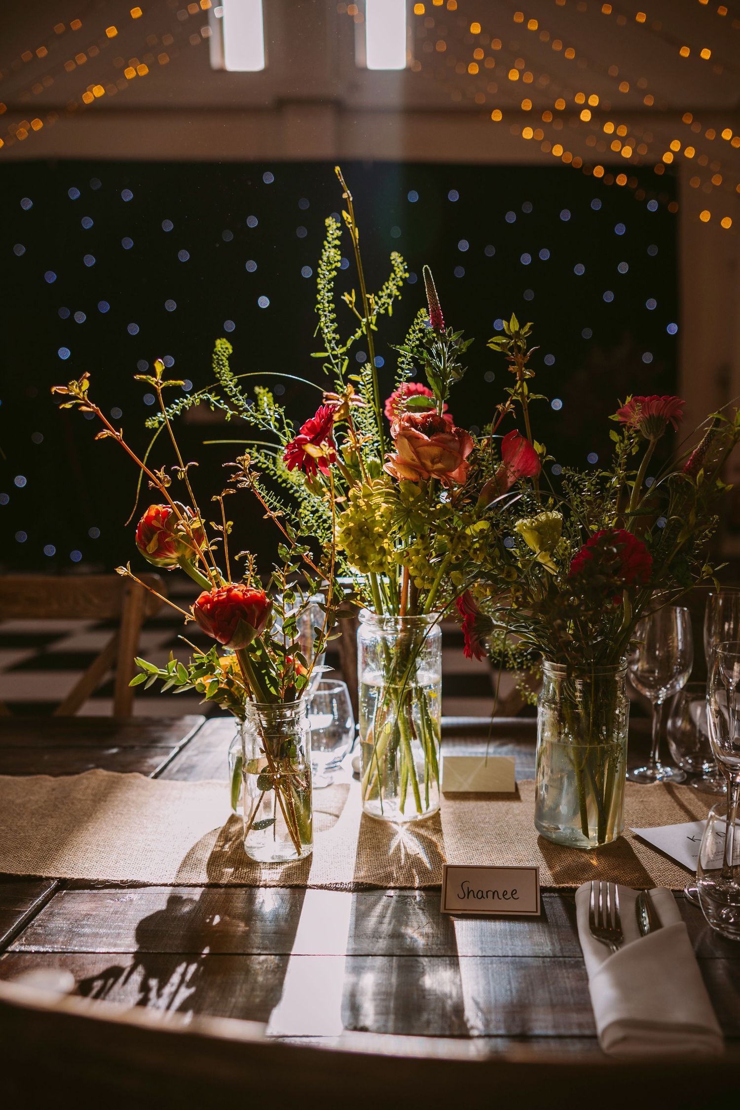 Glass vases with wildflowers and roses on a table setting with string lights glowing in the background.