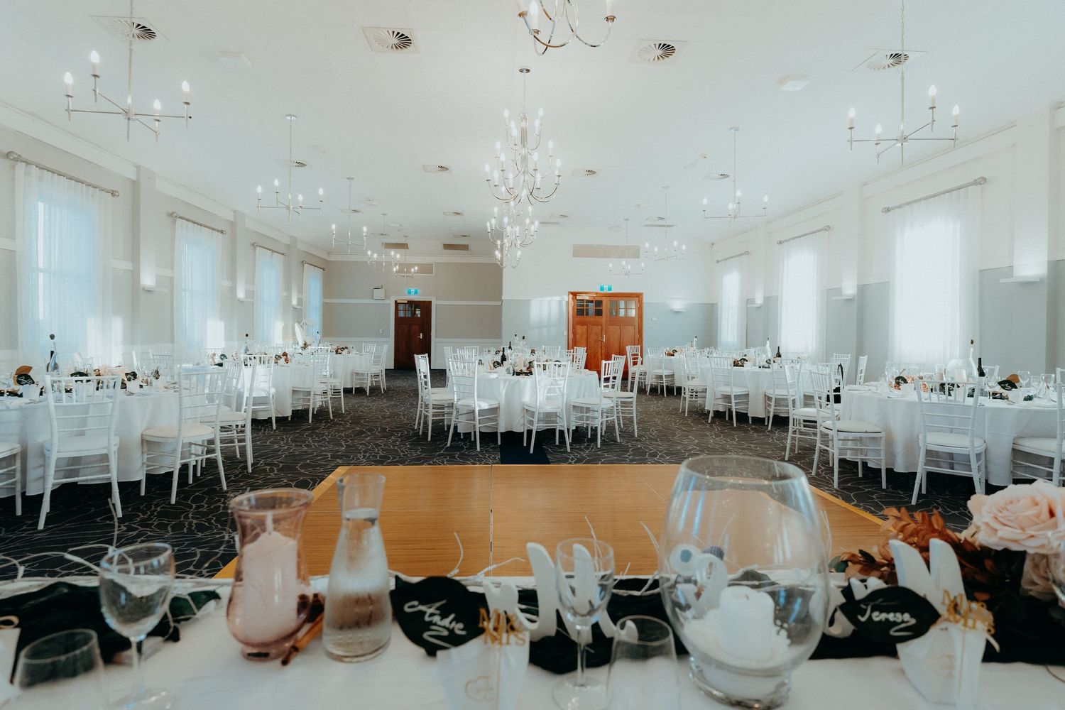 An elegant white reception hall with round tables, crystal chandeliers and a polished wooden dance floor in the center.