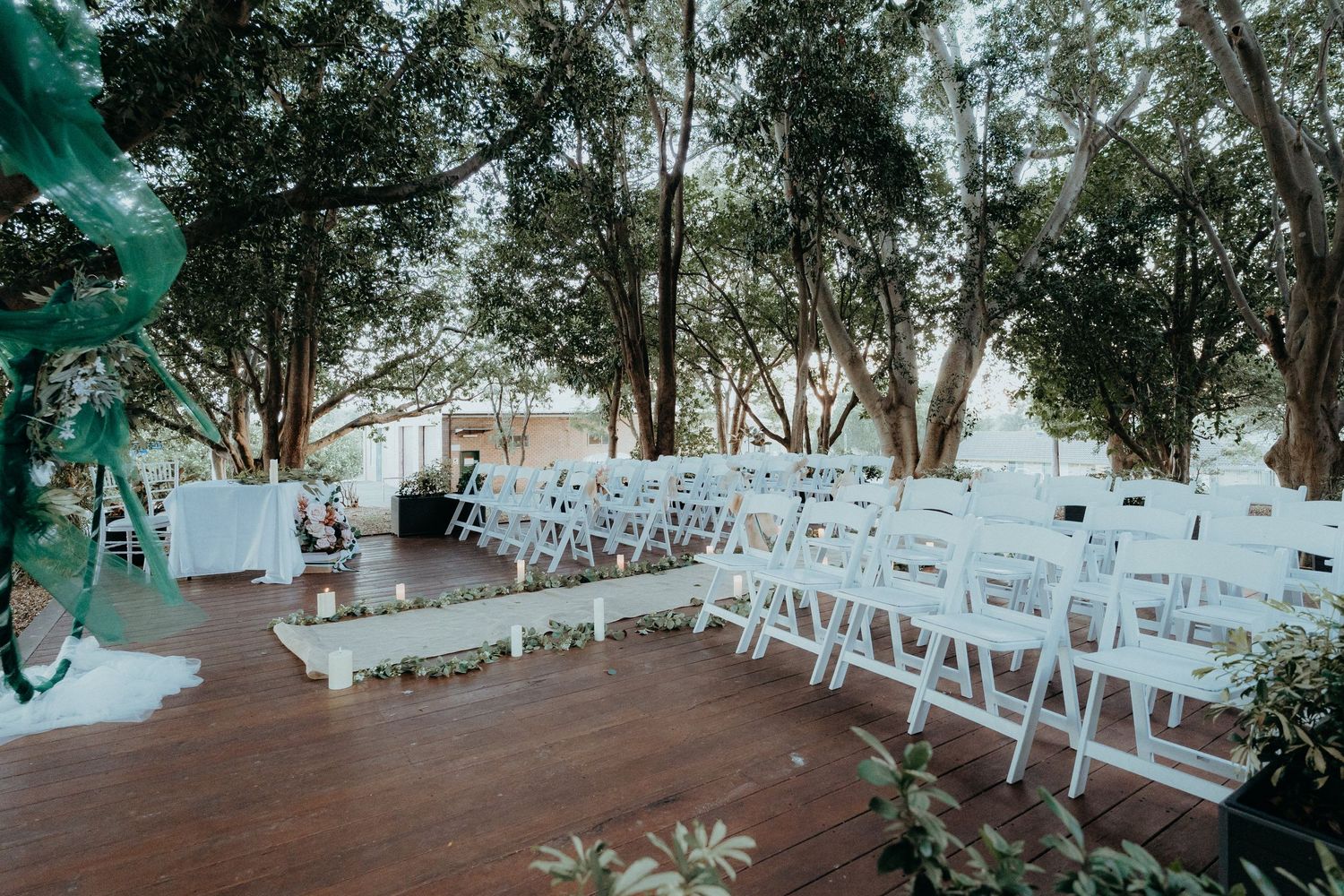 Outdoor wedding ceremony setup with white folding chairs on wooden deck surrounded by trees in natural lighting.