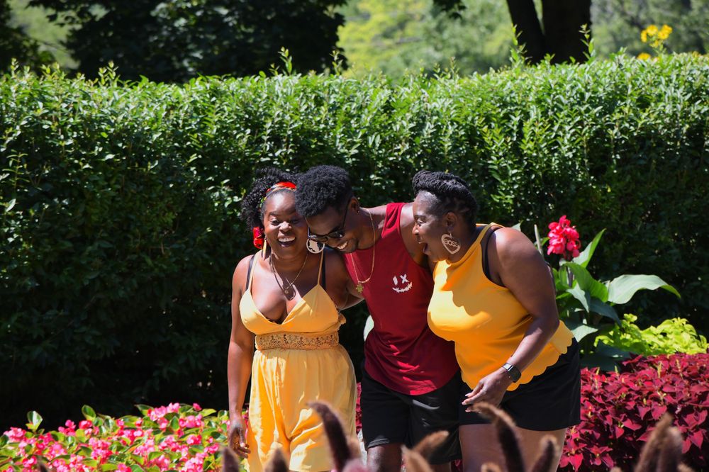 Friends enjoying a sunny day together in a garden with colorful flowers and hedges in the background.