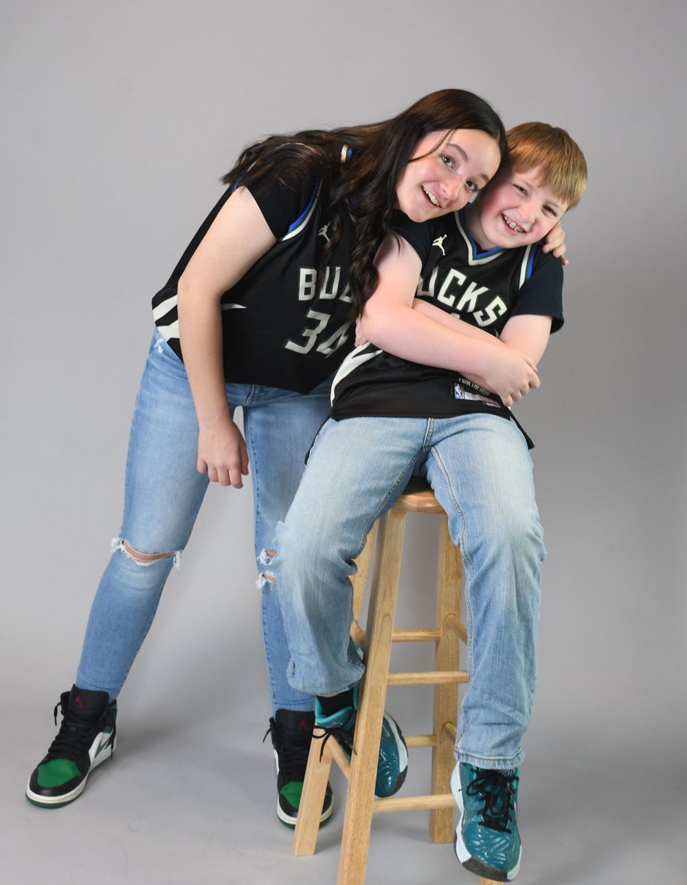 Two people wearing matching black jerseys and ripped jeans lean playfully against a wooden stool in a studio setting.