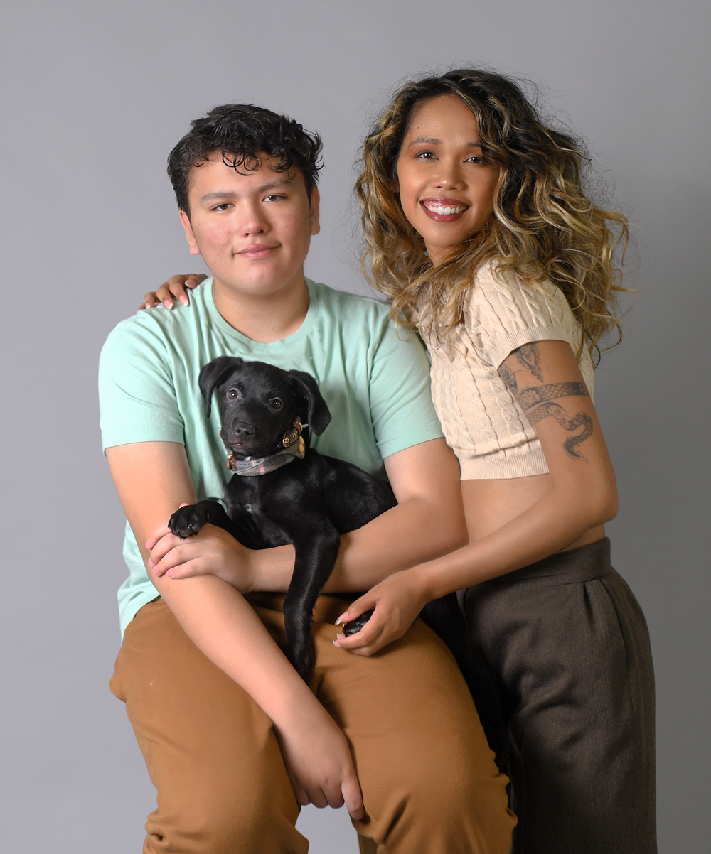 Studio portrait featuring two people and their small black dog against a grey background.