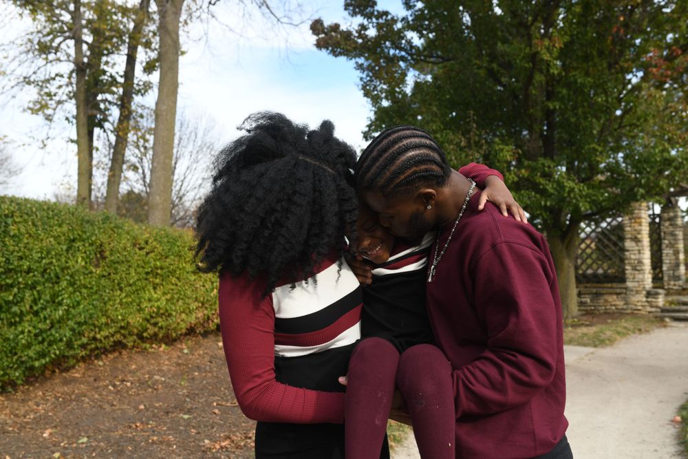 Two people share a tender moment outdoors along a path with green hedges and trees in the background.