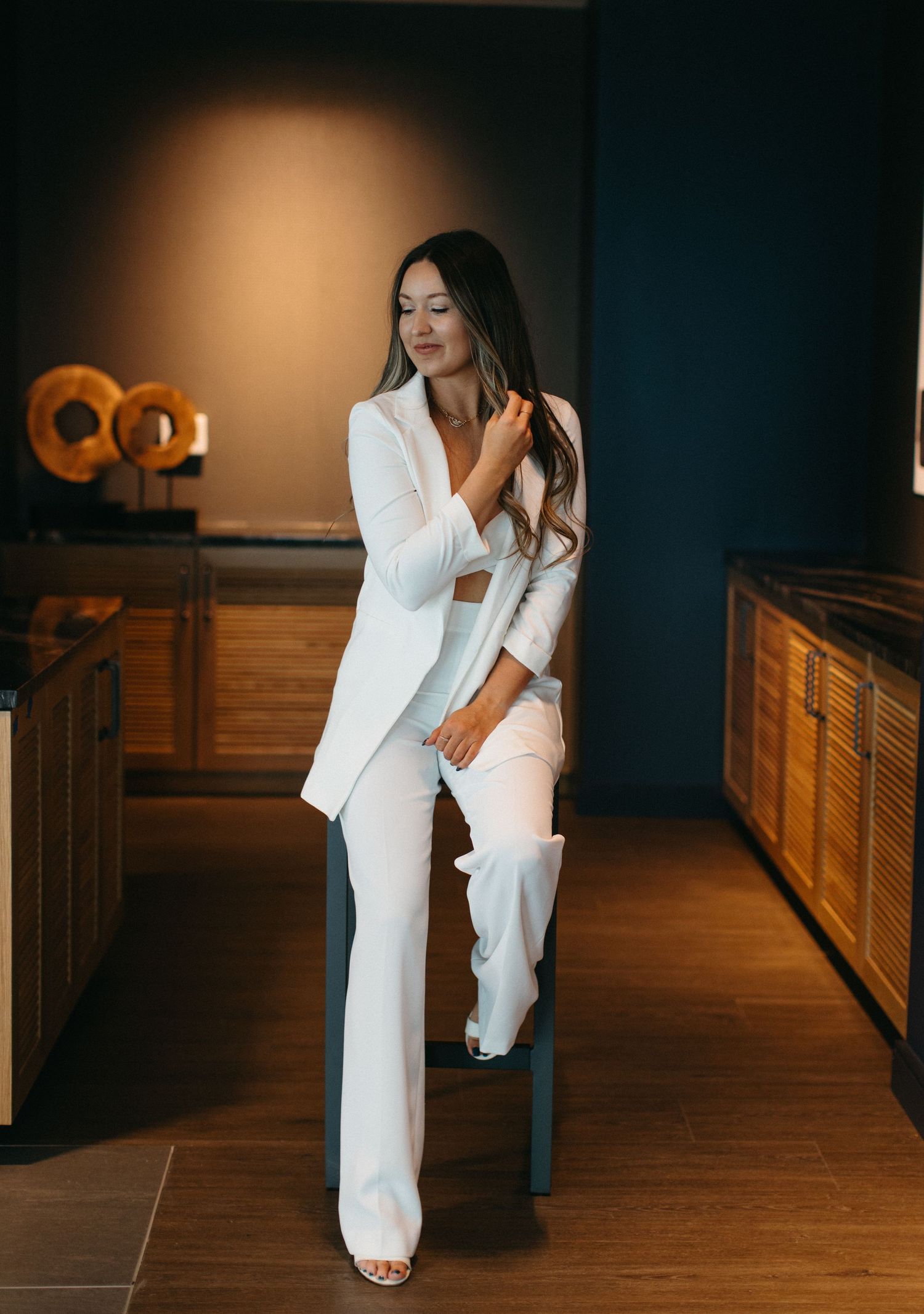 Elegant white pantsuit ensemble paired with high heels on hardwood floor against dark blue walls and wooden cabinetry.