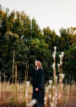 Girl in cap and gown standing in field for senior session photography. Captured by Maryland Photographer Mallory Bramlett.