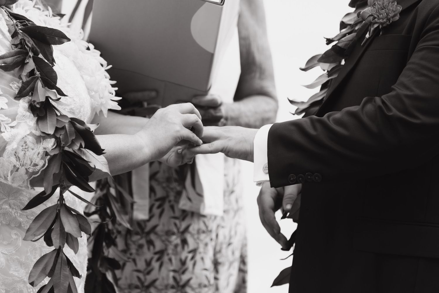 Black and white close-up of hands exchanging rings during a traditional Hawaiian wedding ceremony.