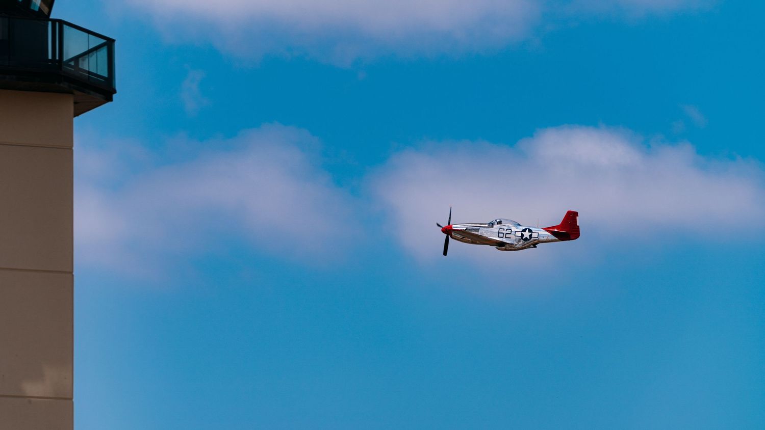 A vintage P-51 Mustang aircraft flying against a blue sky with white clouds.