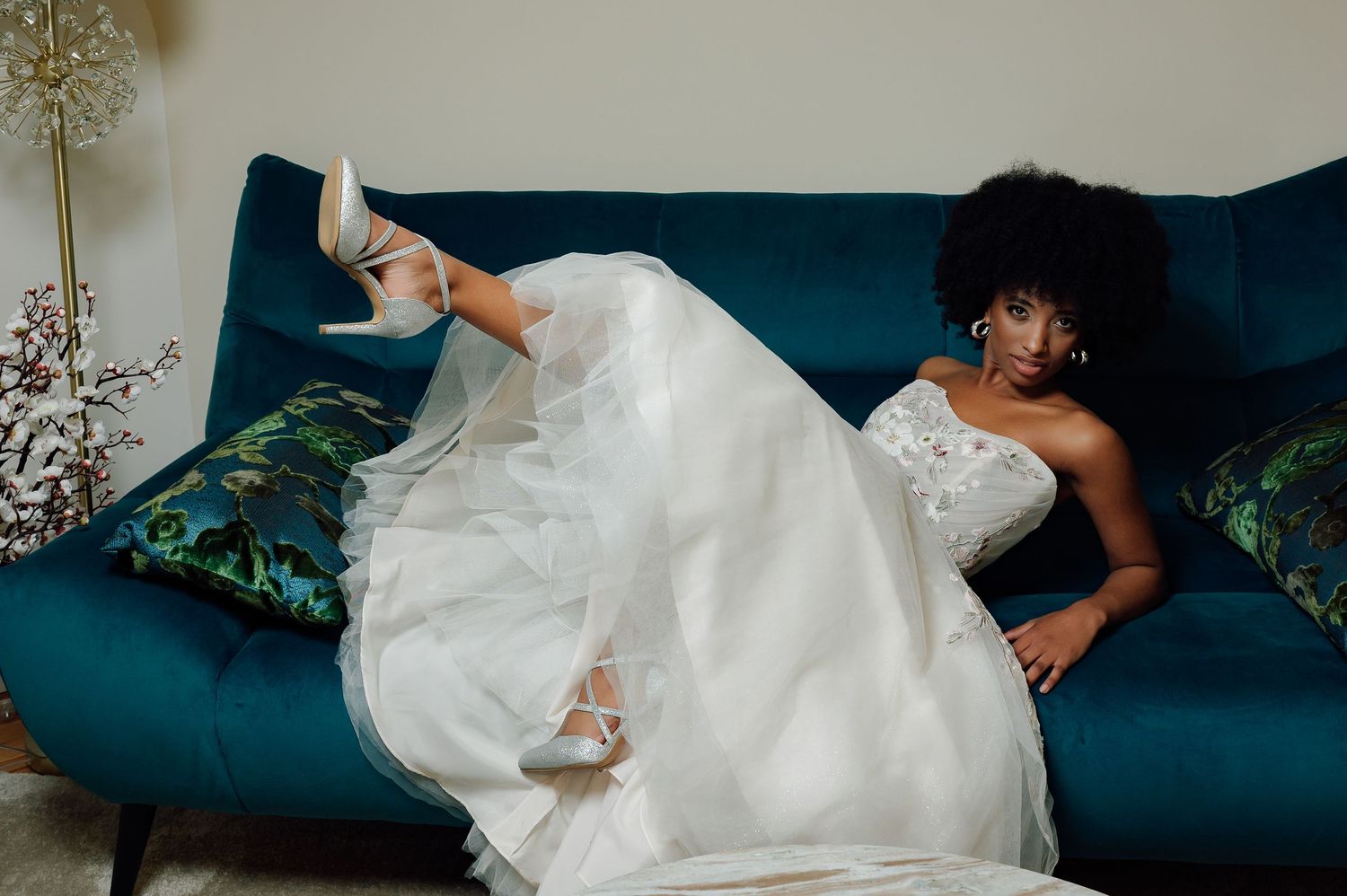 Bride in flowing white gown relaxing on blue velvet couch with decorative pillows.