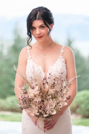 Bride in lace dress holding dried floral bouquet against mountain backdrop.