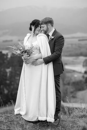 Romantic wedding couple embracing on hillside with scenic mountain views in black and white.