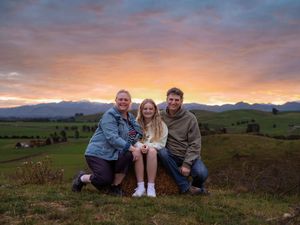 Family of three posing together on a hill at sunset with mountains and green fields in the background.