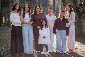 Family of seven posing outdoors in coordinated earth-tone outfits, smiling together in a sunlit wooded setting.