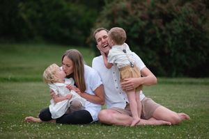 Happy family sitting on grass, parents smiling with two young blonde toddlers during outdoor photo session.