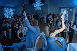Couple raises shoes during lively wedding reception with crowd and snowflake decorations in blue-lit ballroom.