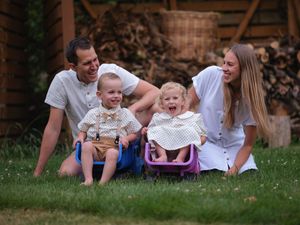 Happy family with two toddlers playing in garden swings, parents smiling on grass with stacked firewood backdrop.
