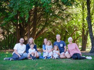 Multi-generational family of eight poses together on grass under large shade trees in casual summer clothing.