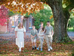 Family of four walking under a large tree in autumn park with colorful fall foliage and brick building backdrop.