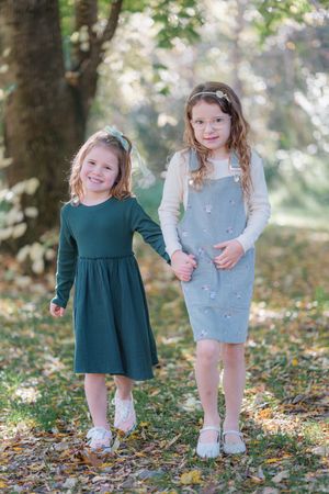 Two young girls in dresses hold hands smiling in an autumn park with fallen leaves and trees.