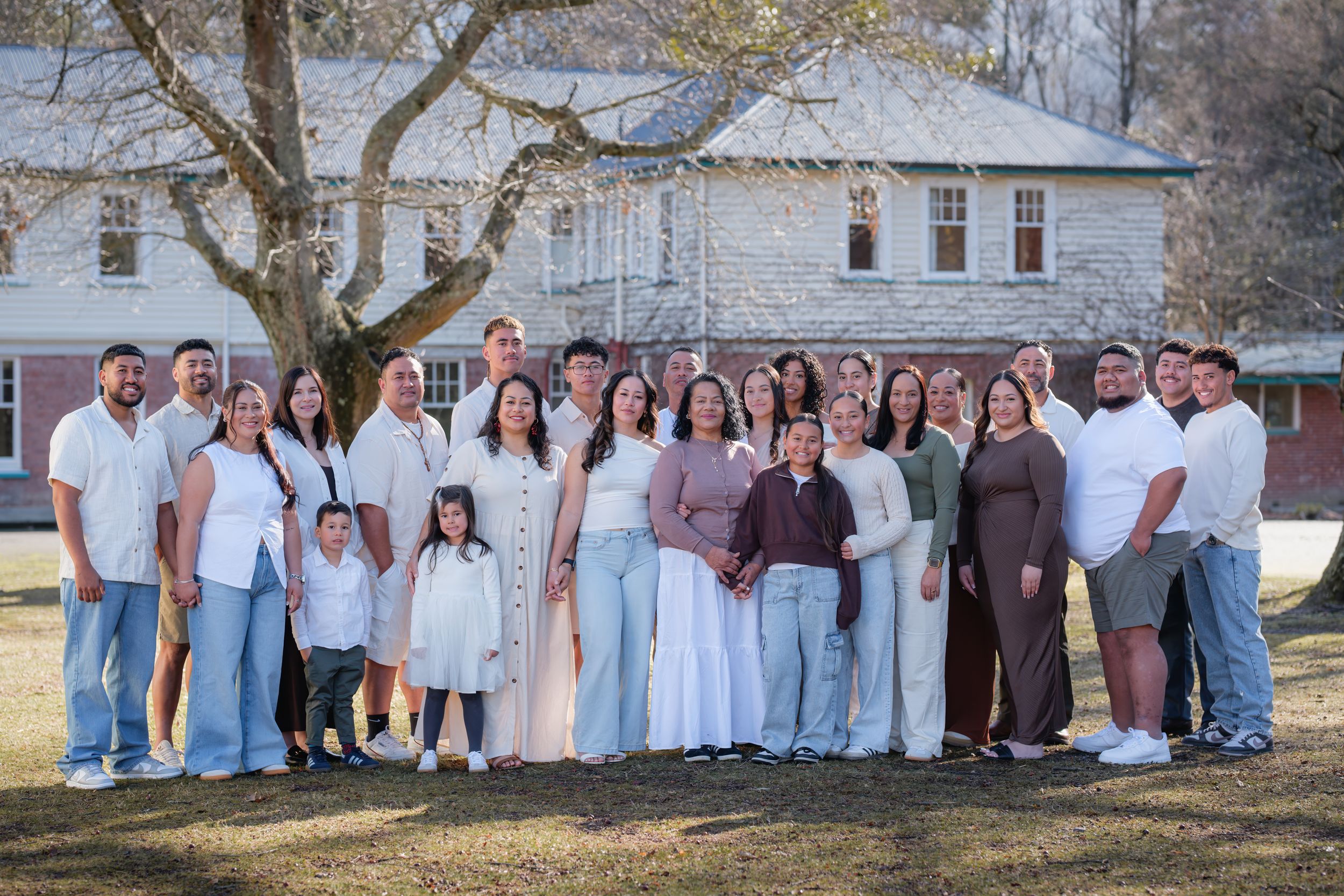 Large multigenerational family posing outdoors near a tree with a white farmhouse in the background on a sunny day.