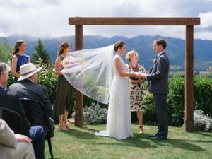 A romantic outdoor wedding ceremony under a wooden pergola with a flowing white veil catching the breeze.