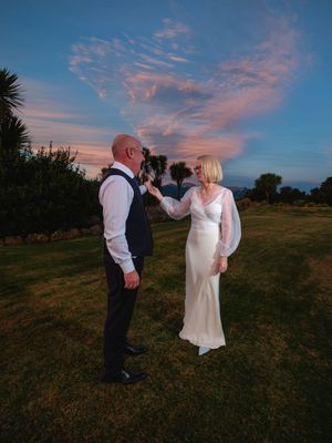 A couple in wedding attire walks hand in hand across a grassy field during a beautiful sunset with pink clouds.
