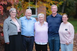 Five adults smiling together outdoors in front of colorful autumn foliage for a family portrait.