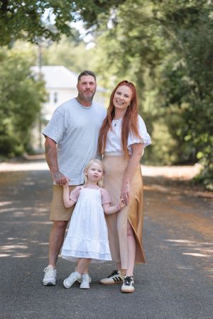 Family of three posing outdoors on a tree-lined path, parents holding hands with young girl in white dress.