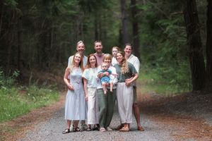 Family of eight poses on a forest gravel path surrounded by tall evergreen trees in coordinated neutral outfits.