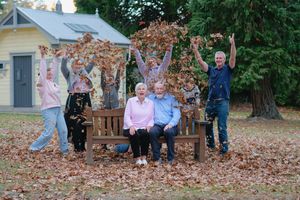 Family group throwing autumn leaves in the air around an elderly couple seated on a wooden bench outdoors.