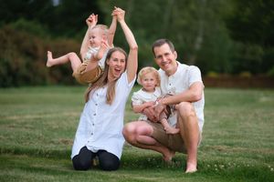 Happy family of four posing outdoors on green grass, parents holding two toddlers, smiling in casual white outfits.