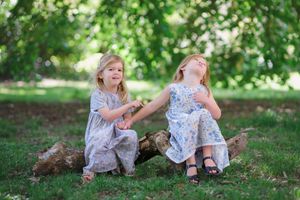 Two young children in floral dresses sit together on grassy ground during a sunny summer day in a park.
