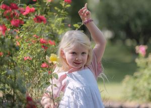 Young blonde girl in white dress pointing up among vibrant red roses in a sunny garden.