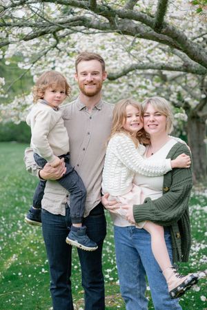 Family of four posing outdoors under a blooming tree during spring, parents holding two young children in casual attire.