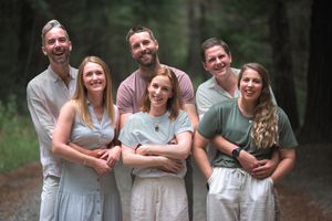 Group of six smiling adults posing together on a forest path surrounded by tall trees.