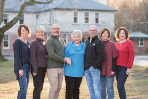 Group of seven adults smiling together outdoors near a brick building during autumn.