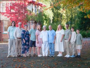 Large multigenerational family posing outdoors in autumn, surrounded by colorful fall foliage near a brick building.