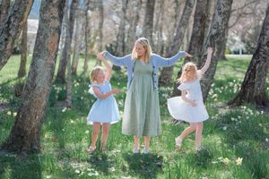A woman in a green dress dances with two young girls in white dresses among spring trees and daffodils.