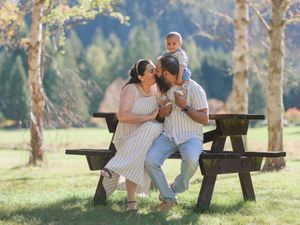 A happy family with a baby sits at a wooden picnic table in a lush green park surrounded by trees.