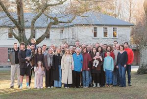 Large multigenerational family group portrait outdoors in front of a white colonial house during autumn.