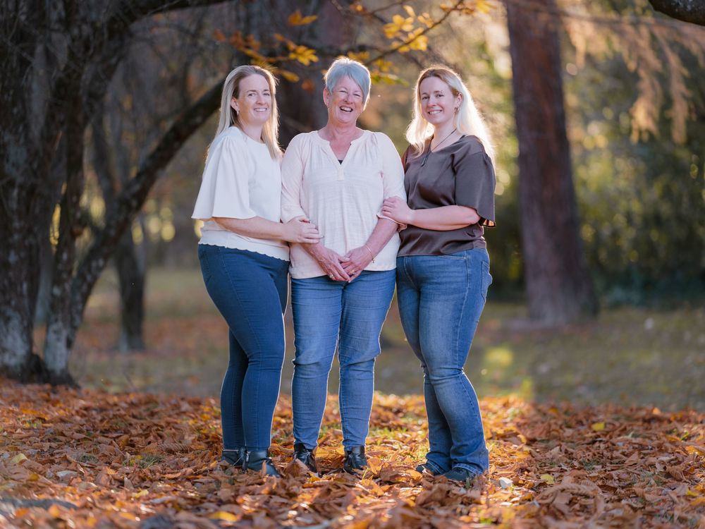 Three women smiling together outdoors among autumn leaves and trees in a warm fall setting.