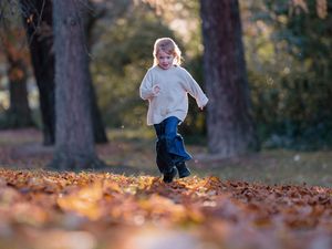 A young child runs joyfully through a park covered in autumn leaves, wearing a white sweater and black boots.