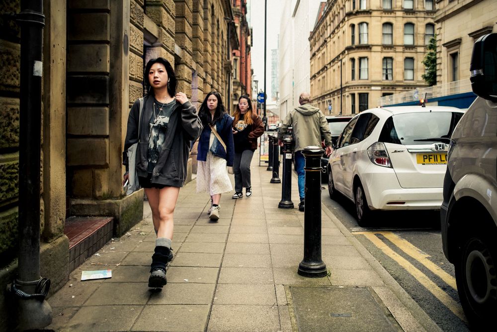 People walking on a busy urban sidewalk alongside historic buildings and parked cars.