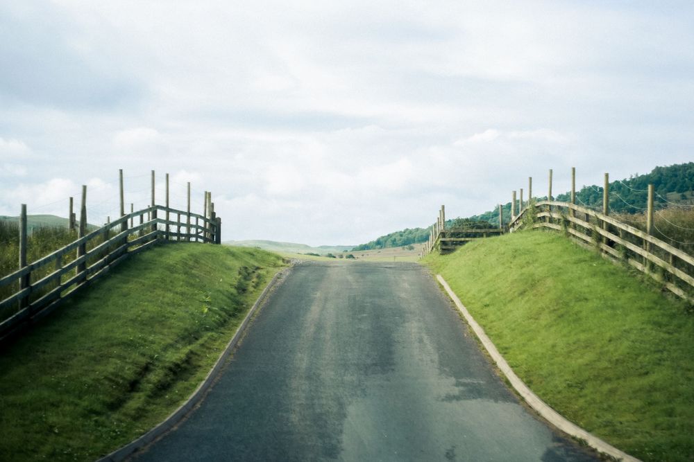 A rural road descending between grassy hills with wooden fences on both sides.