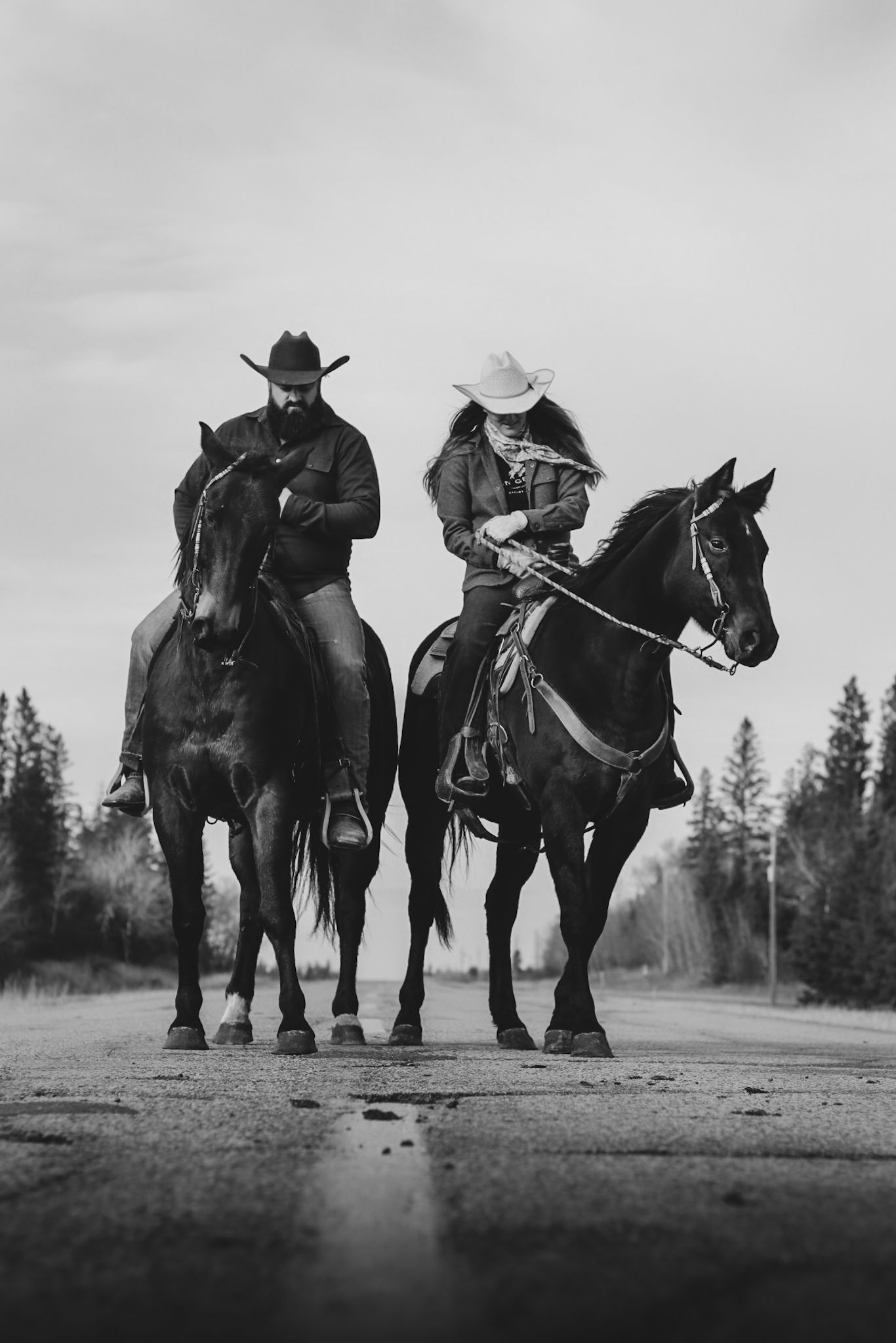 Two equestrians riding dark horses on a wet road in dramatic black and white photography.