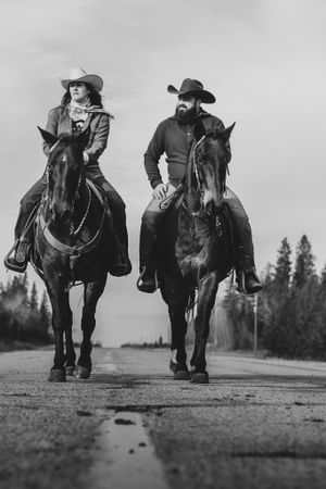 Two riders on horseback in Western attire walking down a rural road in black and white photography.