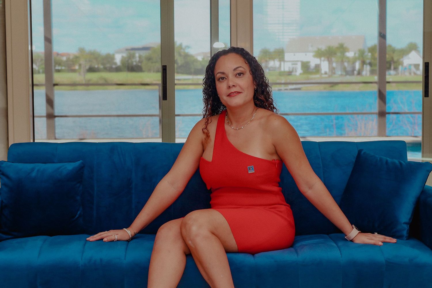 Woman in red dress sitting on blue velvet sofa with waterfront view behind her.