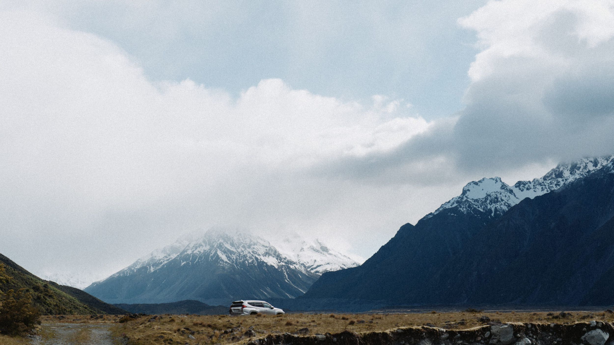 Snow-capped mountains rise behind barren valley under cloudy sky in New Zealand landscape.